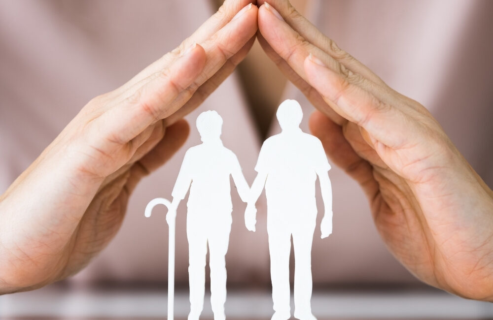 Silhueta branca de um casal de idosos sob a proteção de duas mãos com as pontas dos dedos se tocando